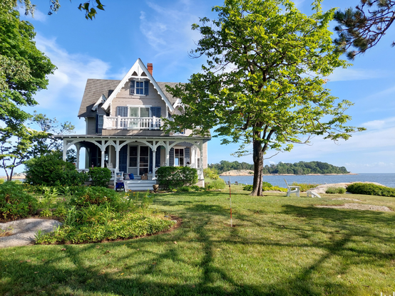 A gable front carpenter Gothic cottage with elaborate white painted trim and cross gable roof, wrap around porch, and Long Island Sound and Atlantic Ocean in the background. The sky is blue with wispy white clouds.
