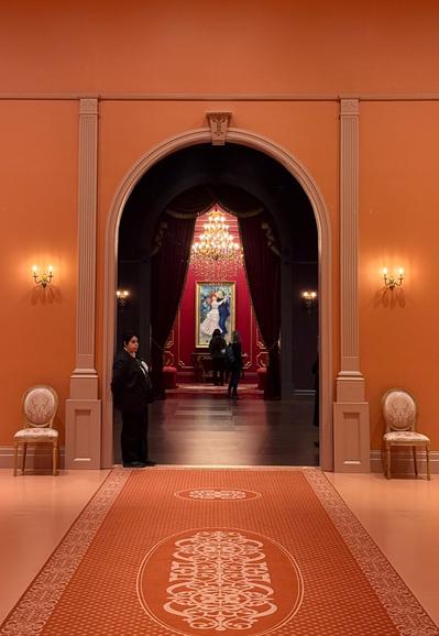 The same Renoir painting sits at the end of a hall. A chandelier sits above it. The round archways of the intervening rooms are draped with curtains. The nearest room is terracotta and has a long carpet and a couple of chairs.