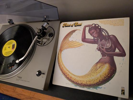 record playing on turntable next to album cover with art of a beautiful black mermaid with braids and pearls on a white background