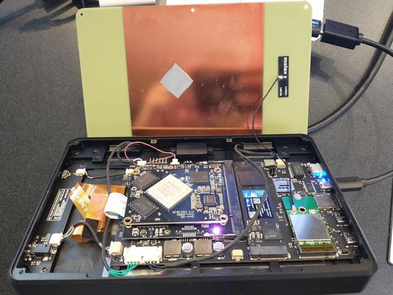 An MNT Pocket Reform open on a lectern.  The motherboard and Rockchip system-on-module board are visible, as well as an M.2 SSD and Wi-Fi adapter, several flat flex and ribbon cables, and various round cables.  The top cover is propped up behind with its shiny copper heat sink and shield layer and a gray patch of thermal tape.  A Micro HDMI cable leaves on the right, presumably heading to the projector.  (It's definitely heading to the projector.)
