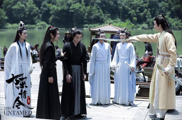 An image is a group standing on a dock consisting of, from viewer's left to right, Lan Wangji, Wei Wuxian, Wen Ning, Lan Sizhui, Lan Jingyi, and Jin Ling. Jin Ling has his sword drawn and extended toward Wen Ning, who exhibits a defeated posture - arms hanging down at his sides, shoulders slumped, and head tilted forward so that his gaze is focused on the ground. Wuxian has his left hand on Wen Ning's arm to provide reassurance while his head is turned to look at Jin Ling. The Lans mostly seem to be simply rendered spectators to the interaction, though Wangji stands near to Wuxian, ever ready to protect the other man as needed.