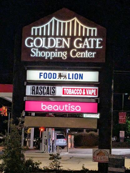 Large tall illuminated stand-up sign in the night with Golden Gate bridge logo, also Food Lion, Rascals, Tobacco & Vape, and beautisa.