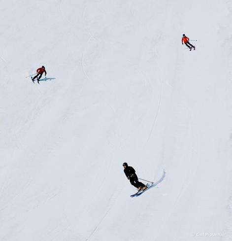 A wide-angle photo of three skiers turning down an open ski slope at Snoqualmie Pass. The snow is smooth and bright, with subtle tracks marking their carved paths. Two skiers wear bright jackets—one red, one orange—while the third is dressed in black. Their spacing and arcs create a pleasing sense of rhythm against the expansive white background, capturing both motion and minimalism.