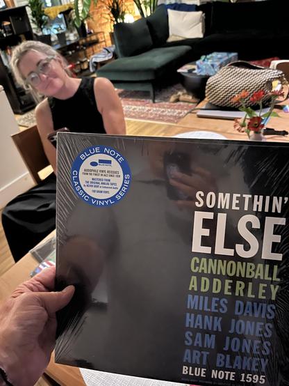 This photo shows someone [me, Claude, me!] holding a vinyl record album in a cozy living room setting. The record is “Somethin’ Else” by Cannonball Adderley, released on the famous Blue Note Records label (catalog number 1595). You can see the distinctive Blue Note circular logo at the top left of the album cover. The album features notable jazz musicians including Miles Davis, Hank Jones, Sam Jones, and Art Blakey.

In the background, there’s a [beautiful] woman with blonde hair wearing a black top, sitting in a comfortable living space with a green couch, coffee table, and various household items. The setting has a warm, homey atmosphere with natural lighting coming through what appears to be windows. [uh… it’s night] 

This is a classic jazz album - “Somethin’ Else” is actually one of the few albums recorded under Cannonball Adderley’s name that features Miles Davis, making it quite a significant recording in jazz history.​​​​​​​​​​​​​​​​