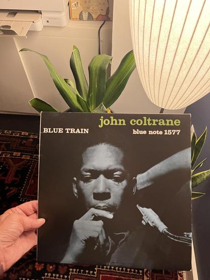 This is a photograph of the iconic “Blue Train” album by John Coltrane, released on Blue Note Records (catalog number 1577). The album cover features a striking photograph of Coltrane in profile, showing him in a contemplative pose with his hand near his face. You can see part of his saxophone in the lower right portion of the image.

The album title “BLUE TRAIN” appears in white text on the left side, while “john coltrane” and “blue note 1577” are displayed in yellow text on the right side against the dark background. This is one of the most recognizable and celebrated jazz album covers, representing a landmark recording from 1957.

The record is being held by someone’s hand [cough cough], and in the background you can see a home setting with a snake plant, a modern pendant light fixture, and some other stuff. There’s also a Persian rug visible in the lower portion of the image.

“Blue Train” is considered one of Coltrane’s masterpieces and a classic of hard bop.​​​​​​​​​​​​​​​​