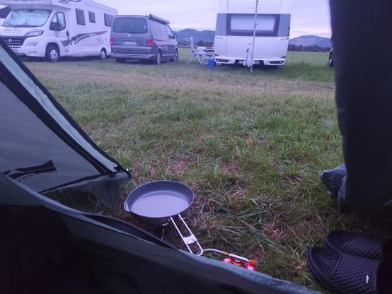 Blick aus Zelt. Im Vordergrund kocht ein Campingkocher Wasser, auf der Wiese stehen Wohnmobile. Wetter ist bedeckt.