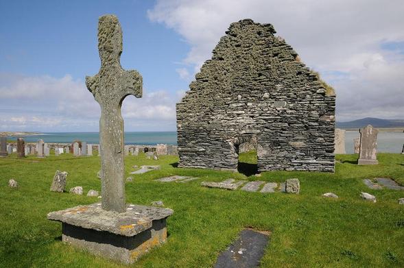 Kilnave Chapel. The image shows a grassy graveyard sloping down to a blue sea in the background. On the left is a badly eroded carved cross standing on a plinth. In the upper right of the frame is the gable end of a stone building with a low door and no windows. The scene is in sunlight.