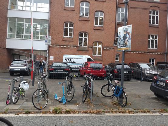 Blick auf ein Backstein-Schulgebäude. Im Vordergrund ein paar Fahrräder und Roller. Daneben an einem Laternenmast ein Plakat. Aufschrift: Vorsicht: Schulanfänger! CDU.