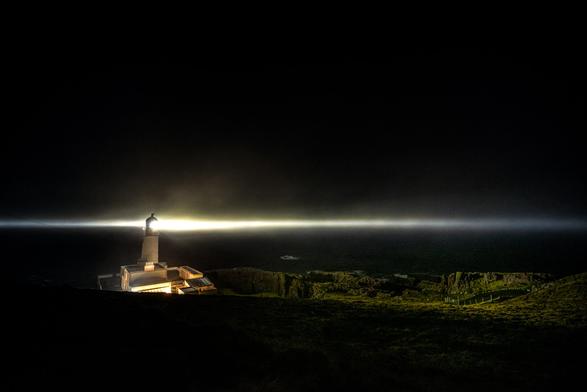 Ein nachts beleuchteter Leuchtturm, der einen hellen Lichtstrahl über das dunkle Meer schickt. Im Vordergrund liegt eine felsige, grüne Landschaft.