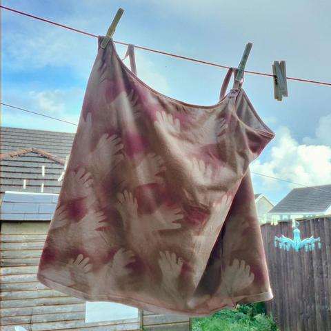 A beige or tan strappy nursing top hangs on a washing line to air out: it's got spray painted hand prints all over it, much like a cave painting.