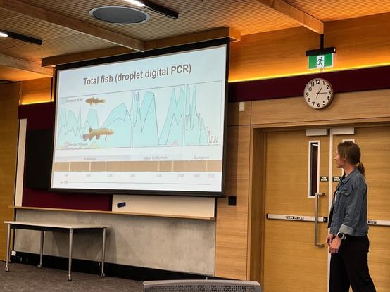Susie Wood stands next to a slide that shows the fish eDNA in sediments of Lake Waihau in Gisborne, which used to be surrounded by forest and is now surrounded by native pasture. Fish DNA collapses in the past 50 years, including the extinction of the common bully, which is a resilient generalist native species. The other fish featured, the banded kōkopu, went extinct in this lake in Māori settlement when the forests were burned off.
