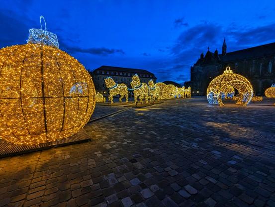 A wide-angle, low-shot of a large public square at dusk, decorated with golden, illuminated Christmas lights. In the foreground, on the left, is a giant, glowing Christmas bauble. To its right are a line of four horse-like figures, also made of golden lights, leading towards lit-up Hemispheres. In the background, a dark cathedral stands against the deep blue sky. The sky is dark blue with clouds. The ground is paved with cobblestones, which are wet and reflective. The overall scene is festive and wintry.