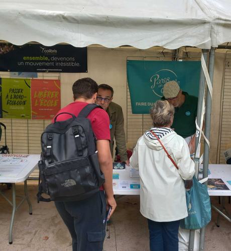 Deux personnes, un homme jeune et une femme d'un certain âge, de dos, en train de discuter avec les bénévoles Paris en Selle 15ème sur le stand du forum des associations du 15ème. En arrière-plan sur le barnum, on voit une bâche aux couleurs de Paris en Selle