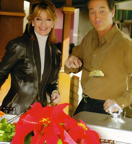 Deidre Hall and Drake Hogestyn stand at a buffet station during a festive indoor event, circa late 2000s. Hall wears a dark leather jacket over a white turtleneck, smiling warmly, while Hogestyn serves food in a casual button-up shirt. A red poinsettia adds holiday flair to the scene
