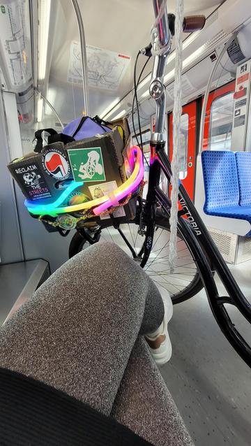 Hochkant in Farbe. Blick von der fotografierenden Person aus auf sitzende Beine in Glitzerstrumpfhosen und den vorderen Teil eines schwarzen Hollandrades. Vorne ist ein schwarzer Pappkarton als Korb angebracht, darauf verschiedene antifaschistische Sticker. Um den Korb leuchtet ein Lichterschlauch in Regenbogenfarben. Das Rad steht in einer s Bahn.