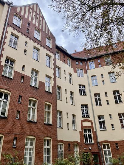 Rückseite eines Wohnhauses mit Balkonen, hellem Putz, rotem Klinker im Erdgeschoss und umgeben von Bäumen.