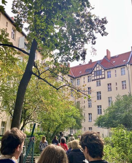 Rückseite eines Wohnhauses mit Balkonen, hellem Putz, rotem Klinker im Erdgeschoss und umgeben von Bäumen.