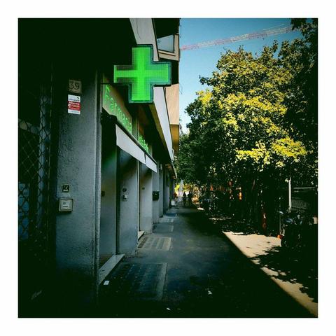 Farbfoto, aufgenommen heute in einer Straße in Rom. In der Mitte schwarz das asphaltierte Trottoir. Rechts grüne Bäume. Links Geschäftsgebäude. Oben grün leuchtend das Symbol einer Apotheke. Mobilfotografie mit Vignette for Android auf Xiaomi.