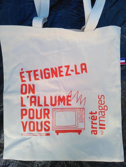 Un sac avec marqué dessus "éteignez-la on l'allume pour vous" avec un dessin de télé et une allumette, le logo d'arrêt sur images est marqué sir le côté