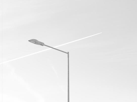 Streetlight with a sleek, modern design against a pale sky. A diagonal contrail runs behind it, creating a contrasting line across the sky. Minimalist composition with a monochromatic color scheme.