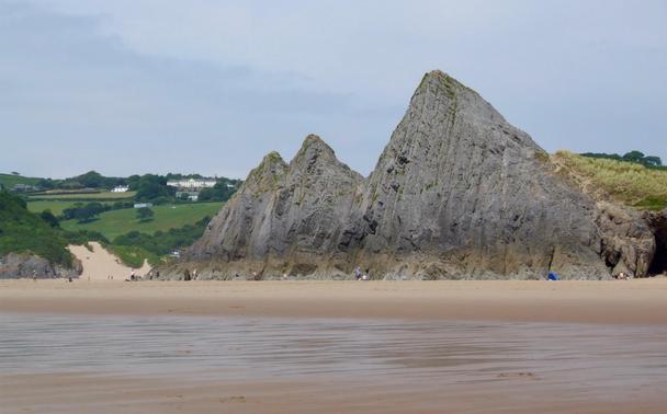 On the sweeping sands of Three Cliffs Bay in Swansea, Wales, dramatic limestone peaks rise sharply from the golden beach, forming a striking natural gateway to the sea. The tide has retreated, leaving rippled patterns across the wet sand, while the green hills and dunes in the background create a serene contrast to the rugged cliffs. Small figures of visitors appear scattered along the shore, emphasizing the scale of the towering rock formations. Beyond the cliffs, the gently rolling countryside is dotted with white farmhouses and clusters of trees, completing the idyllic coastal scene that defines the Gower Peninsula, an Area of Outstanding Natural Beauty.