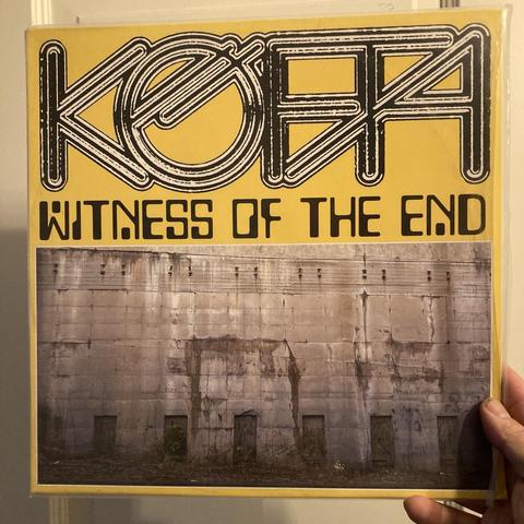 Holding a copy of the album with my right hand. Artwork base color is yellow, with layout design split roughly 50/50 vertically.
Top half contains the artist & album title, using two sort of retrofuturistic fonts. Bottom half has a sepia tone picture of perhaps a old, huge concrete wall with some wet spots on it. The wall has seven doorway like shapes to the ground edge of it