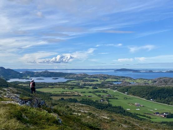 View from the mountain towards the coast where small islands are scattered. A wide green valley with farms and farmland leads towards the coast. A man is standing in the distance looking into the horizon. A sunny day with white scattered clouds.