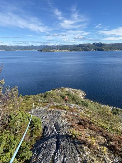 From the downward climb. A blue rope for support is fastened to the rugged and rocky trail leading downwards to the fjord. A man can be seen in the distance walking down along the rope.