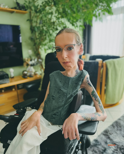 Ich sitze selbstbewusst, aber entspannt in meinem Wohnzimmer und trage ein graugrünes, ärmelloses Oberteil und eine helle Hose. Meine Arme sind mit bunten Tattoos bedeckt. Mit meinem leichten Lächeln wirke ich zufrieden und bereit für ein Date. Im Hintergrund sind helle Vorhänge, Holz­möbel und eine große grüne Pflanze zu sehen, die dem Raum eine warme Atmosphäre verleihen.