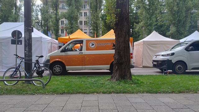 Das Bild zeigt eine Szene in einer Parkanlage mit einer orangefarbenen Van, die mit dem Logo "PIRATENPARTEI" versehen ist. Der Van steht auf einer Rasenfläche vor einem Baum, neben einem geparkten Fahrrad an einem Fahrradständer. Im Hintergrund sind mehrere Zelte in verschiedenen Farben, darunter Weiß und Orange, aufgestellt. Einige Zelte sind mit Fahnen geschmückt. Im Hintergrund ist ein Gebäude mit mehreren Fenstern zu sehen, und die Umgebung ist von Bäumen umgeben. Die Szene wirkt ruhig und friedlich, mit einem gepflegten Grün und einer gepflasterten Straße im Vordergrund.