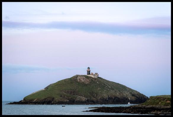 Ballycotton Lighthouse perched on top of a rounded green island with rocky coastline, white lighthouse tower with red band, adjacent buildings, and small boat visible in calm blue waters under a slightly cloudy sky in Co Cork.