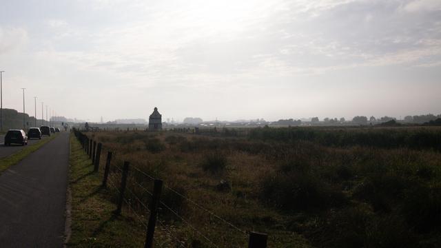 Color photo of a narrow road with a bike path alongside a grassy field under a hazy, overcast sky, with several cars parking between the road and the bike lane. In the distance, a solitary small building stands amid the open landscape, silhouetted against the soft morning light. Blankenberghe is almost visible in the mist at the horizon.
