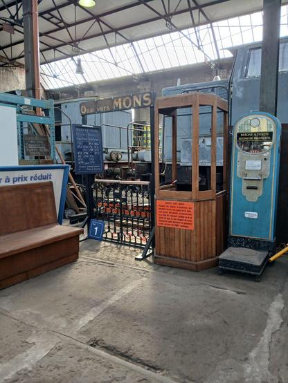 La guérite d'accès aux quais avec le panneau d'affichage à gauche des départs des trains, un banc de la salle d'attente et le pèse-personne à tickets