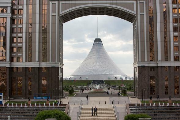 View of the Khan Shatyr Entertainment Center in Astana, Kazakhstan, framed symmetrically by two tall buildings with reflective glass facades. The tent-like structure stands prominently in the center, seen through a large archway. In the foreground, landscaped paths, fountains, and people add scale and vibrancy to the urban scene.