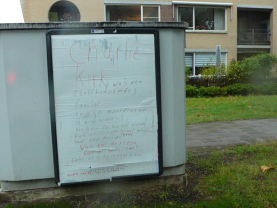 witte poster met handgeschreven op reclame bord op straatkastje in berm voor voetpad met strook groen voor woningcomplex