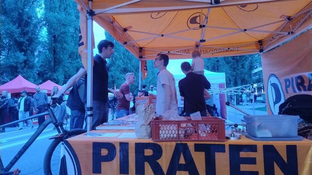 Das Bild zeigt eine Szene an einem Markt oder einer Veranstaltung, bei der ein Stand mit einem großen orangefarbenen Zelt aufgebaut ist. Auf dem Tisch des Stands ist ein orangefarbenes Tuch mit der Aufschrift "PIRATEN" in großen schwarzen Buchstaben ausgebreitet. Auf dem Tisch stehen verschiedene Gegenstände, darunter ein Korb mit Papieren und ein Plastikbehälter. Einige Personen stehen unter dem Zelt und interagieren miteinander. Ein Mann in einem weißen Hemd steht am Tisch und scheint mit jemandem zu sprechen. Ein weiterer Mann in einem schwarzen Pullover hält ein Kind auf dem Arm. Im Hintergrund sind weitere Zelte und Menschen zu sehen, die sich auf der Veranstaltung aufhalten. Ein Fahrrad steht neben dem Stand. Die Atmosphäre wirkt entspannt und freundlich.