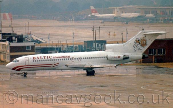 Side view of a 3 engined jet airliner, with the engines mounted on the rear fuselage, taxiing from right to left at a damp and dismal airport.
The plane is mostly white, with a pair of red stripes on the lower forward fuselage, with red "Baltic International" titles on the upper forward fuselage.
The red registration "YL-BAE" is on the engine pods mounted on the sides of the rear fuselage, complete with a small Latvian flag.
The tail has a black and white line drawing of a rampant lion with a golden mane, holding a golden shield outlined in white, with a white letter "B" in the middle, and 3 golden stars at the top.
Wet, grey concrete apron fills the foreground, with a couple of brick buildings stretching across the middle of the frame in the background.
There are more buildings in the distance on the right of the frame, with a couple of cargo planes parked outside, with trees filling the distance on the left, under dreary grey sky.