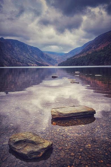 A serene, eye-level shot of a lake in Glendalough, Ireland, under a cloudy, dramatic sky. The surface of the water reflects the sky and the surrounding mountains. In the foreground, the water is clear, revealing large, smooth rocks on the lake bed, and a large piece of driftwood rests partially submerged. A waterfall is visible in the distance between the mountains. The mountains are a mix of dark greens and purples, giving the scene a moody, yet tranquil feel.