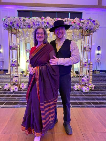 Two people stand in front of the wedding canopy, which is covered in flowers. The woman has long salt and pepper hair and is wearing a burgundy sari edged in red and gold with a red blouse and red and gold high heels. Her watch shows through the pallu. The goateed man has a broad-brim black hat, black vest, and black pants with an undyed shirt.