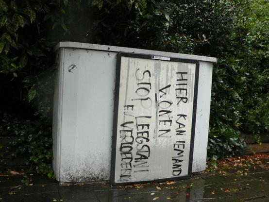 kastje voor groene begroeiing met wit bord in zwart om zn kant geschreven