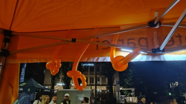 Das Bild zeigt eine Szene unter einem orangefarbenen Zelt, das mit Luftballons geschmückt ist. Die Ballons sind in verschiedenen Formen geformt, darunter Knoten und Schlaufen. Im Hintergrund ist ein Gebäude mit dem Schild "Partners" zu sehen, und es scheint sich um eine städtische Umgebung zu handeln, da man auch andere Zelte und Menschen erkennen kann. Die Szene ist bei Nacht aufgenommen, was durch die Beleuchtung und die Dunkelheit im Hintergrund deutlich wird. Die Atmosphäre wirkt lebendig und gesellig, möglicherweise auf einer Veranstaltung oder im Rahmen einer öffentlichen Aktivität.