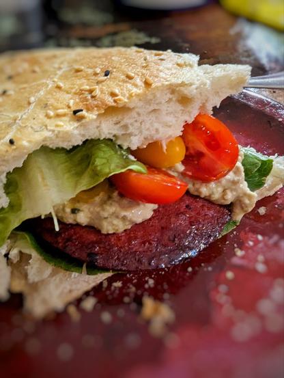 Dunkelroter Teller mit einer Ecke knusprigen Fladenbrots darauf. Das Brot ist gefüllt mit einer großen Scheibe Sucuk, Tomatenhälften, Salat.