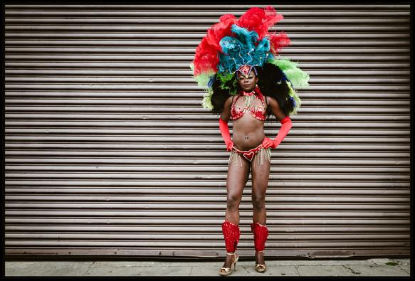 A portrait of dazzling woman in a Carnaval outfit - red gloves, leg warmers, and bikini... with a giant feathered headdress. She stands on the sidewalk, hands on hips, and casts a strong gaze at the camera. Behind her - a corrugated garage door makes strong horizontal strips leading the eye in the woman's direction.