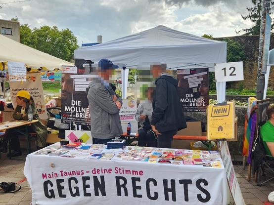 drei Personen stehen unter einem Pavillon. Vor ihnen ein Tisch mit Aufklebern und Informationsmaterial. Der Tisch ist beschriftet mit MTK GEGEN RECHTS