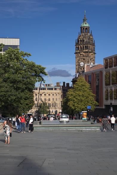 This photograph shows a sunny day at Barkers Pool in Sheffield, a public square dominated by the city's historic Town Hall clock tower on the right. The tower, with its distinctive green spire, rises against a clear blue sky.
In the foreground, the paved square is sparsely populated with a few people walking in different directions. 
To the left, a large, mature tree with a full canopy of green leaves partially frames a light-coloured, traditional building. On the opposite side, a modern building with an intriguing facade of vertical garden panels stands next to the town hall, highlighting a blend of old and new architecture