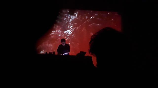 stoi facet przy stole z rozstawionym sprzętem elektronicznym i kręci gałkami, w tle wizualizacje z jakimiś liśćmi zabarwionymi na czerwono, jakaś ludzka głowa trochę zasłania widok