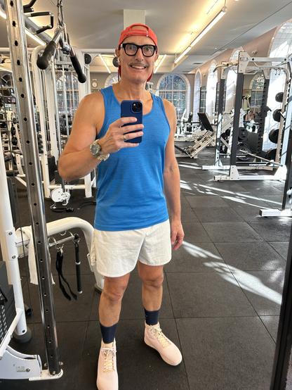 A man (me) poses for a selfie in a gym. He wears a blue tank top, white shorts, and light pink shoes with navy socks. A red baseball cap is worn backward. The gym equipment is visible in the background.