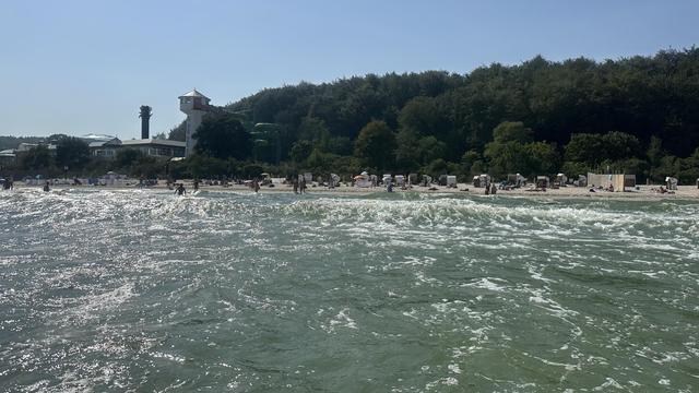 The image shows a view of a sandy beach from the water. In the foreground, gentle waves can be seen, their surface reflecting the sunlight.
The beach stretches horizontally across the centre of the photo, where people, both clothed and naked, are walking along the water's edge or relaxing naked on the sand. Several beach chairs and parasols are placed along the coast. The nudist area of the beach is located between the two wooden fences; outside the fences, swimwear is mandatory.
Behind the beach rises a wooded hill densely covered with green trees. On the left are buildings with white facades, one of which has a tower-like structure and a green water slide winding down it. The sky above is clear and blue, with no visible clouds. The entire scene depicts a typical summer day in a coastal town, with natural and recreational elements.