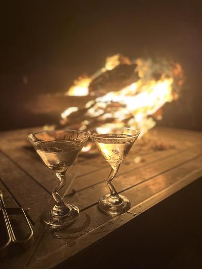 The image features two martini glasses filled with a clear gin martini, positioned on a wooden surface. The glasses are slightly tilted, with condensation visible on their surfaces, suggesting a cool beverage. The background is dominated by a large, bright fire, with flames and logs visible, creating a warm and inviting atmosphere. The fire's glow casts a soft light on the scene, enhancing the ambiance. To the left of the glasses, a pair of metal tongs is partially visible, adding to the setting's rustic charm. The overall composition highlights the contrast between the coolness of the drinks and the warmth of the fire, creating a cozy and intimate setting.
Provided by @altbot, generated privately and locally using Ovis2-8B
🌱 Energy used: 0.166 Wh