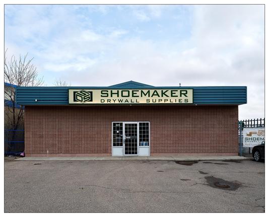 Single-story commercial building with a large sign reading "SHOEMAKER DRYWALL SUPPLIES" on the facade. The building has a brick exterior, a blue roof, and a central entrance with double glass doors. There's a small, leafless tree to the left and a blue gate enclosing a side area to the left of the structure. The sky is overcast and there are puddles on the concrete parking area in front.